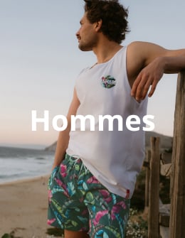 Homme pr&egrave;s de la plage se tenant contre une cl&ocirc;ture avec une planche de surf dans les mains, portant une camisole et un maillot de bain de la marque Hamabe.