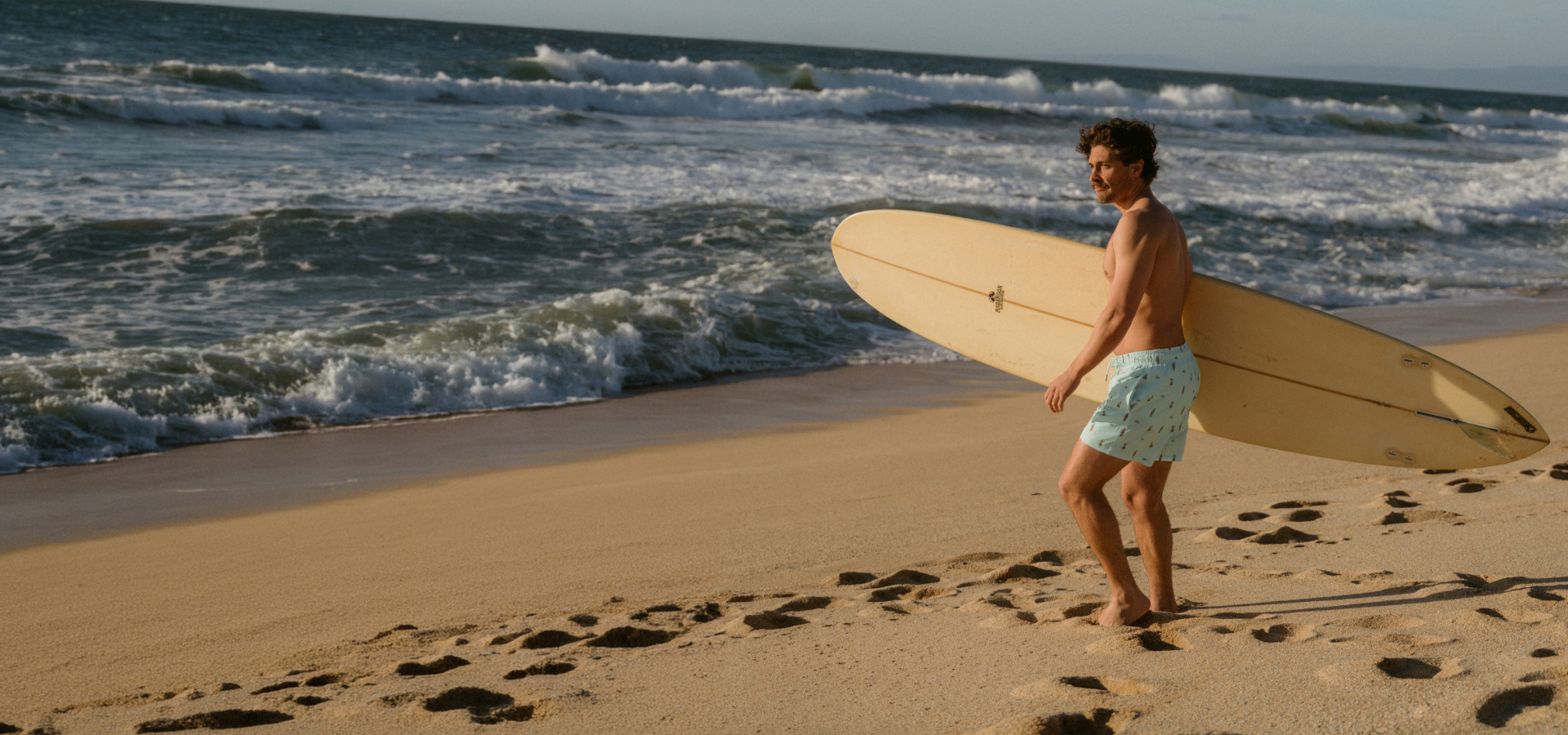 A man on the beach holding a surfboard in his hands and wearing Hamabe swim trunks.
