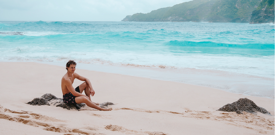 Man sitting on a beach and wearing black swim trunks from the brand Hamabe.